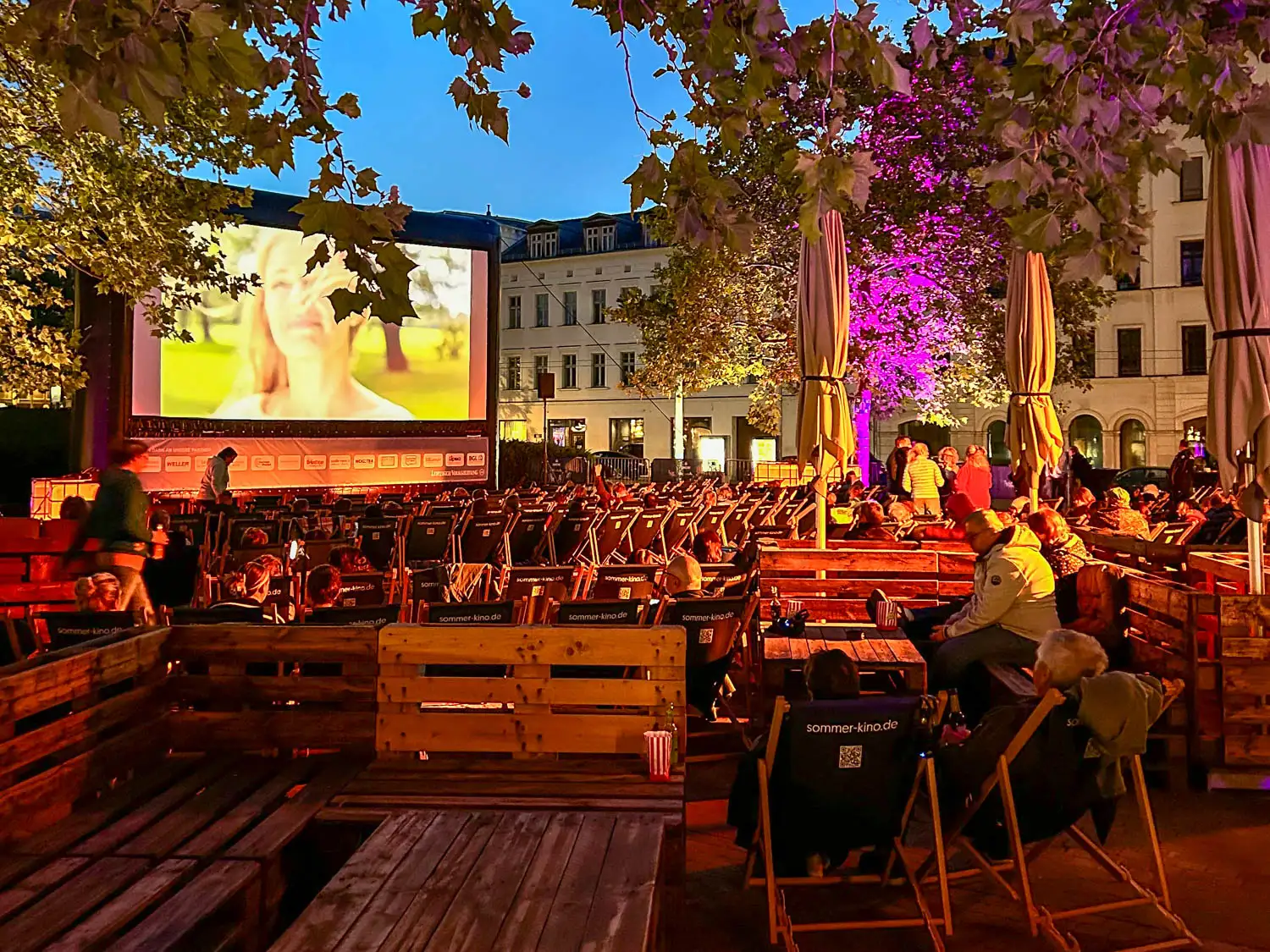 Abendliches Open-Air-Kino mit Sitzgelegenheiten aus Holzbänken und Liegestühlen. Vorne sitzen Besucher entspannt mit Snacks, während auf der großen Leinwand ein Film läuft. Bäume mit violetter Beleuchtung und Häuserfassaden umrahmen die stimmungsvolle Sommerkinoszene.