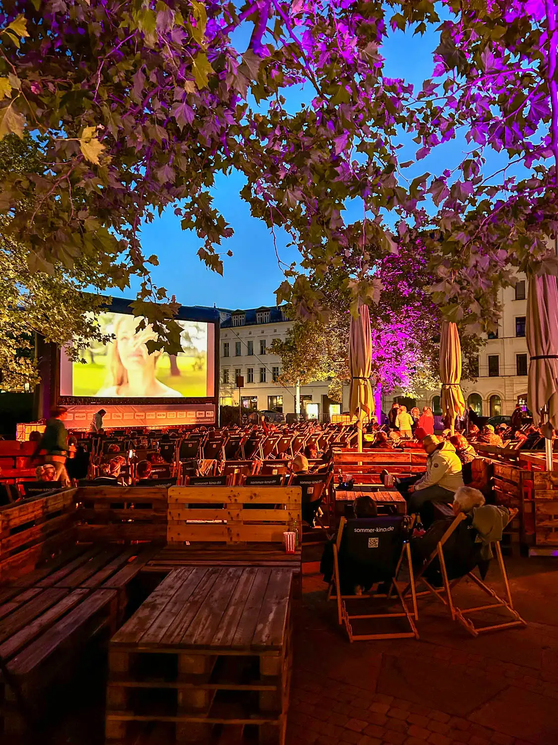 Abendliches Open-Air-Kino mit Sitzgelegenheiten aus Holzbänken und Liegestühlen. Vorne sitzen Besucher entspannt mit Snacks, während auf der großen Leinwand ein Film läuft. Bäume mit violetter Beleuchtung und Häuserfassaden umrahmen die stimmungsvolle Sommerkinoszene.