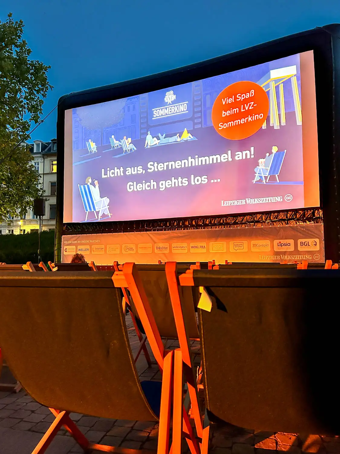Open-Air-Kino mit aufgestellten Liegestühlen vor einer großen Leinwand am Abendhimmel. Auf der Leinwand steht: ‚Licht aus, Sternenhimmel an! Gleich geht’s los … Viel Spaß beim LVZ-Sommerkino‘. Im Vordergrund sind leere Liegestühle zu sehen.