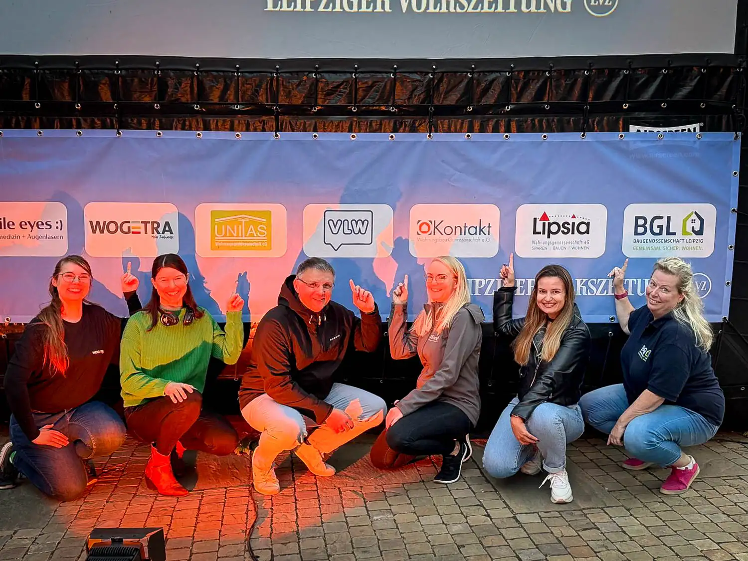 Sechs Personen knien und hocken lächelnd vor einer Sponsorenwand beim LVZ-Sommerkino und zeigen mit dem Finger auf die Logos ihrer Wohnungsunternehmen.